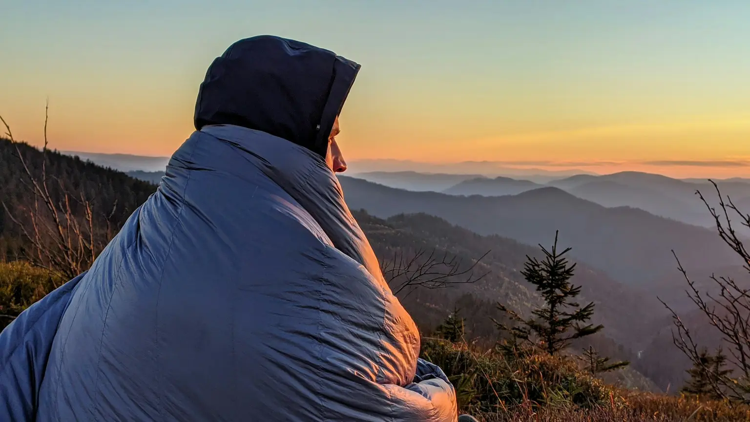 朝焼けの山並みを前に、寝袋にくるまって座る人